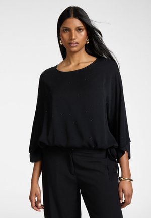 Kobieta o długich czarnych włosach nosi luźną czarną bluzkę z delikatnymi zdobieniami, czarne spodnie, złote kolczyki-koła oraz złotą bransoletkę.