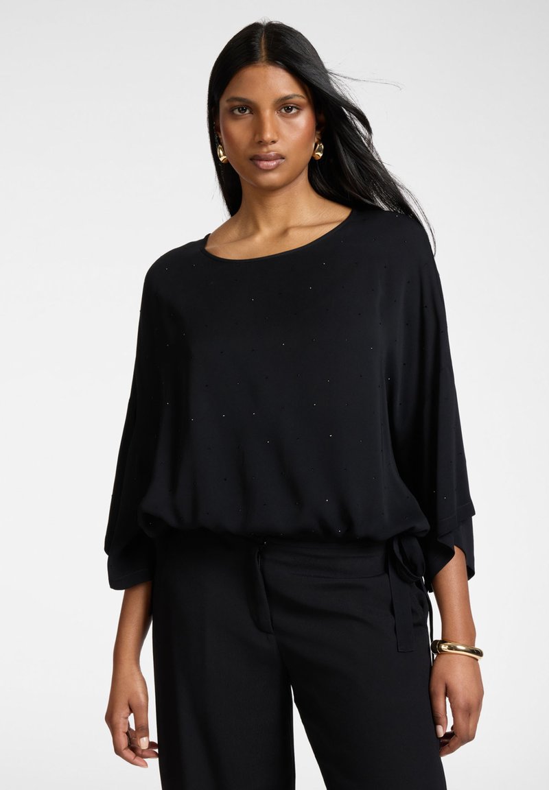 Mujer con cabello largo y negro que lleva una blusa negra suelta con adornos sutiles, pantalones negros, pendientes de aro dorados y una pulsera dorada.