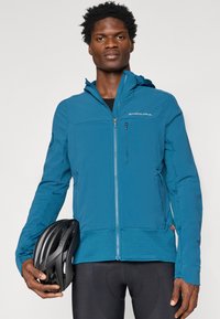 Jachetă de ciclism de culoare turquoise, cu închidere cu fermoar, glugă și mâneci texturate. Include o cască de bicicletă neagră, ținută în mână.