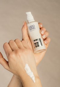 DADO SENS Ectoin Anti-Aging-Fluid in einer transparenten Flasche mit Pumpe, cremige weiße Textur auf dem Handgelenk aufgetragen. Die Haut erscheint glatt.