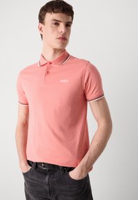 Chemise polo rose avec un col classique, dotée d'une bordure rayée blanche et marron. Portée avec un jean foncé, affichant un logo minimaliste sur la poitrine.
