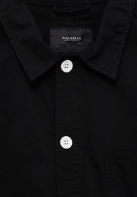 Schwarzes Baumwollhemd mit spitzem Kragen, weißen Knöpfen und einer Brusttasche. Das Etikett trägt "PULL&BEAR". Glatte Textur mit einem klassischen Design.