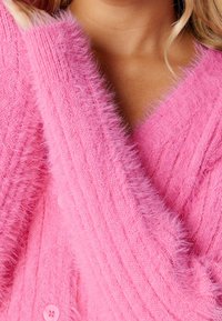 Pink fluffy cardigan featuring textured ribbing, a deep V-neck, and front buttons, with prominent soft fur-like details.