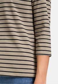 Beiger Baumwolloberteil mit schwarzen horizontalen Streifen, Dreiviertelärmeln und abgerundetem Saum. Der Stoff wirkt weich mit leichter Textur.