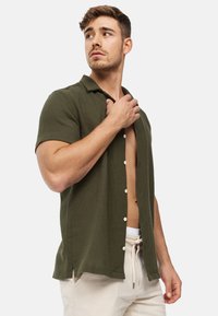 Camicia verde oliva a maniche corte con bottoni, in tessuto testurizzato e colletto, con bottoni bianchi e fessure laterali, indossata aperta sopra pantaloni beige.