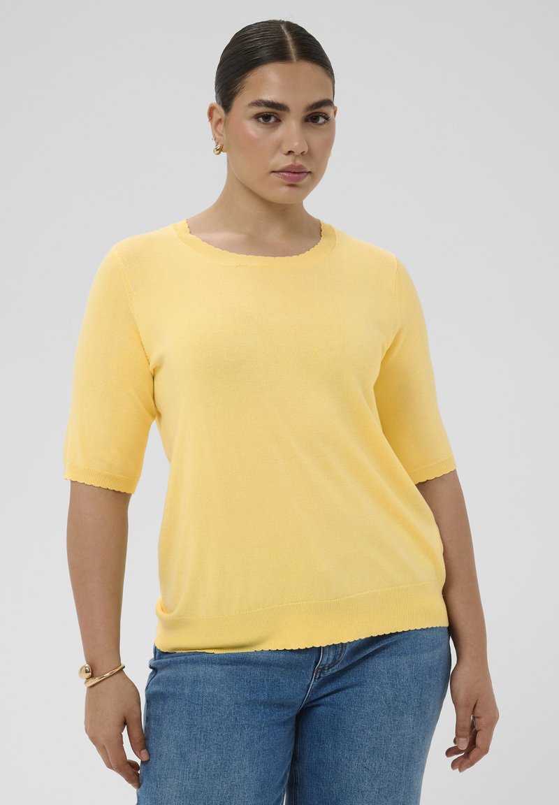Femme aux cheveux foncés en chignon bas portant des boucles d’oreilles créoles en or, un pull jaune à manches courtes, un jean bleu et un bracelet en or, debout devant un fond gris.