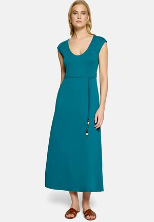 Femme portant une robe longue sans manches bleu sarcelle avec un décolleté arrondi, une fine ceinture à perles à la taille, et des sandales plates marron.