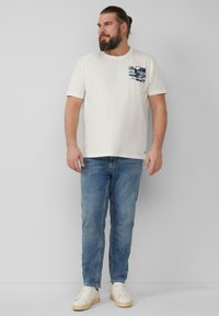 White cotton t-shirt with a navy graphic pocket design, paired with light blue jeans and white sneakers. Plain background.