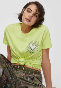 Neonowo żółta koszulka z nadrukiem serca i detalami promieni słonecznych na wysokości klatki piersiowej, zawiązana z przodu. W połączeniu z brązowymi, wzorzystymi spodniami.