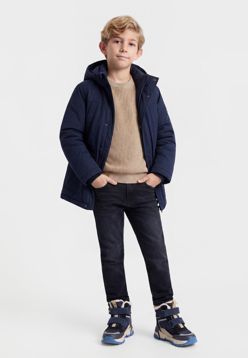 Dreng iført navy hættejakke, beige sweater, sorte jeans og blå vinterstøvler, stående med hænderne i lommerne, neutral baggrund.