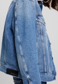 Giacca di jeans blu chiaro con design cropped, bottoni in metallo e un'etichetta sul fondo. Struttura rigida con dettagli di cucitura visibili.