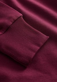 Burgunderfarbener Pulloverkasten mit gerippter Textur; glattes Gewebe mit zartem Glanz, das die tiefen Farben und die detaillierte Naht betont.