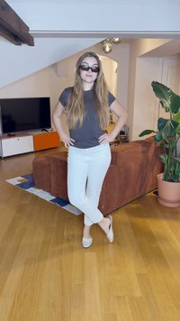 Modellen står i ett vardagsrum iförd en grå t-shirt, vita byxor och ljusgrå ballerina skor. Långt hår och mörka solglasögon; brun soffa i bakgrunden.