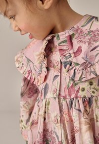 Květinové šaty z růžové látky s volánovým límcem, zapínáním na knoflíky a plisovanou sukní s různými květinami v jemných barvách.