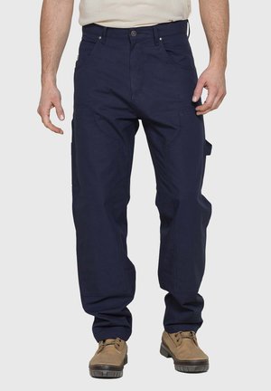 Uomo con pantaloni cargo blu navy e stivali da lavoro color cuoio, in piedi davanti a uno sfondo semplice.
