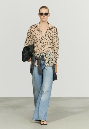 Femme portant un chemisier transparent à imprimé léopard, un jean large bleu clair, des lunettes de soleil noires, tenant une pochette noire tissée, et un pull gris noué autour de la taille.