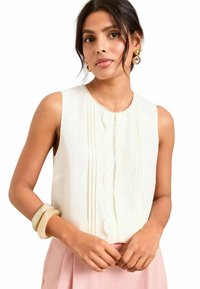 Sleeveless cream blouse with scalloped trim and pleated detailing. Worn with a pink skirt and accessorized with a chunky light-colored bangle.