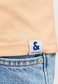 Ourlet de chemise couleur pêche avec une étiquette de marque Jack & Jones blanche au-dessus de la poche d'un jean denim bleu clair et d'un rivet métallique.