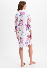 Bloemige linnen jurk met een witte achtergrond, voorzien van roze en blauwe rozenpatronen, driekwartmouwen en zijsplitten. G gedragen met roze sandalen.