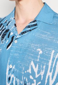 Niebieska koszula z krótkim rękawem z abstrakcyjnym czarno-białym wzorem. Posiada spiczasty kołnierzyk i białe guziki. Gładka, lekka tkanina.