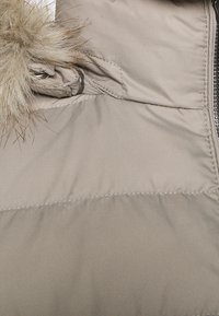 Manteau d'hiver beige matelassé avec une capuche bordée de fourrure et une fermeture éclair argentée visible sur le côté droit.