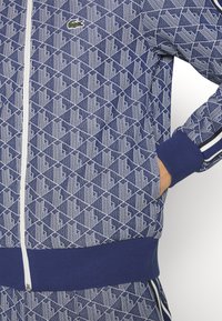 Veste de survêtement à motif géométrique bleu et blanc avec fermeture éclair blanche, poignets et ourlet côtelés, et petit logo crocodile vert sur la poitrine.
