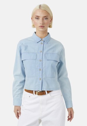 Femme aux cheveux blonds courts portant une chemise bleu clair boutonnée avec des poches poitrine, un pantalon blanc et une ceinture marron.