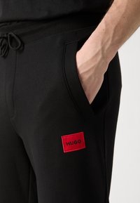 Mano en el bolsillo de pantalones de chándal negros con un parche rectangular rojo con el logotipo "HUGO" en el muslo, usados con una cinturilla negra y cordón.