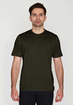 Uomo con capelli scuri corti che indossa una maglietta tinta unita verde scuro a maniche corte con girocollo, in piedi davanti a uno sfondo chiaro, rivolto verso l'osservatore.