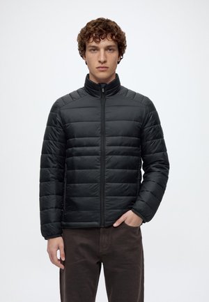 Jeune homme aux cheveux bouclés portant une veste matelassée noire et un pantalon marron, debout avec une main dans la poche sur un fond blanc.