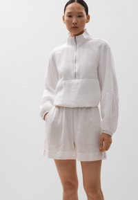 Weißer Leinenpullover mit halbem Reißverschluss und hohem Kragen, kombiniert mit passenden, lockeren Shorts. Mit weit geschnittenen Ärmeln und einer Fronttasche.