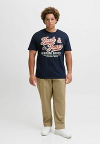Marineblå bomulds t-shirt med hvid og rød grafisk tekst; parret med beige bukser og hvide sneakers; afslappet pasform og behageligt design.
