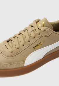 Beige sportsko lavet af ruskind og læder, med et hvidt stribe-design, guld logo accenter og en brun gummisål.