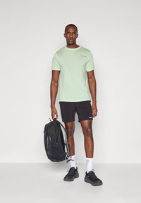 T-shirt de manga curta verde claro com um pequeno logótipo, combinado com calções pretos. A pessoa segura uma mochila preta e usa ténis pretos.