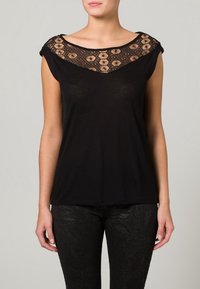 Lyhythihainen musta paita, jossa on pitsiyksityiskohta kaula-aukossa ja kukkakuosi, rento istuvuus, yhdistettynä teksturoituihin mustiin housuihin.