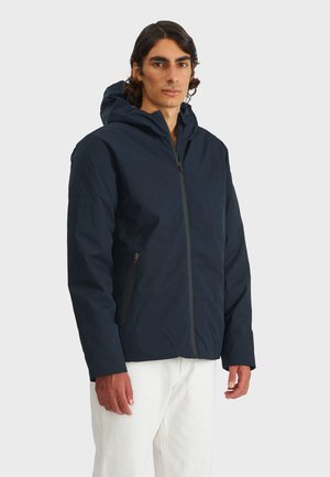 Giacca con cappuccio blu navy con zip frontale, tasche laterali e una texture liscia. Indossata sopra pantaloni bianchi, che mostrano un design casual.
