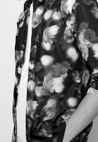 Camisa negra con estampado floral y mangas cortas, con un cierre de botones en la parte delantera y una camiseta blanca que es parcialmente visible debajo.