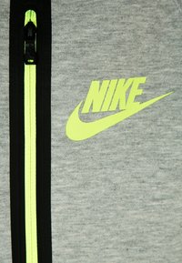 Grijze sweatvest met een rits, voorzien van een zwarte rand en een felgeel Nike-logo. De stof is gestructureerd en geeft een casual, sportieve uitstraling.