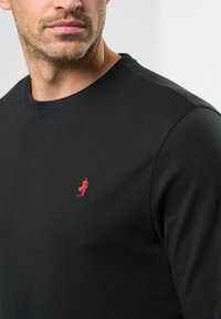 Uomo che indossa una maglietta nera a girocollo con un piccolo logo ricamato rosso sul lato sinistro del petto, visibile dalla collana fino alla parte superiore del torso.