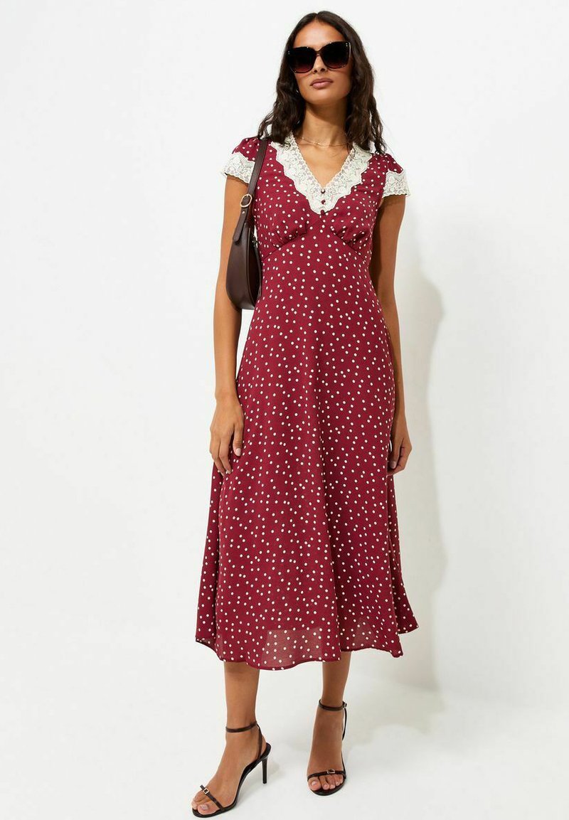 Woman wearing a red polka dot midi dress with white lace trim, black sunglasses, black high-heeled sandals, and carrying a brown shoulder bag.
