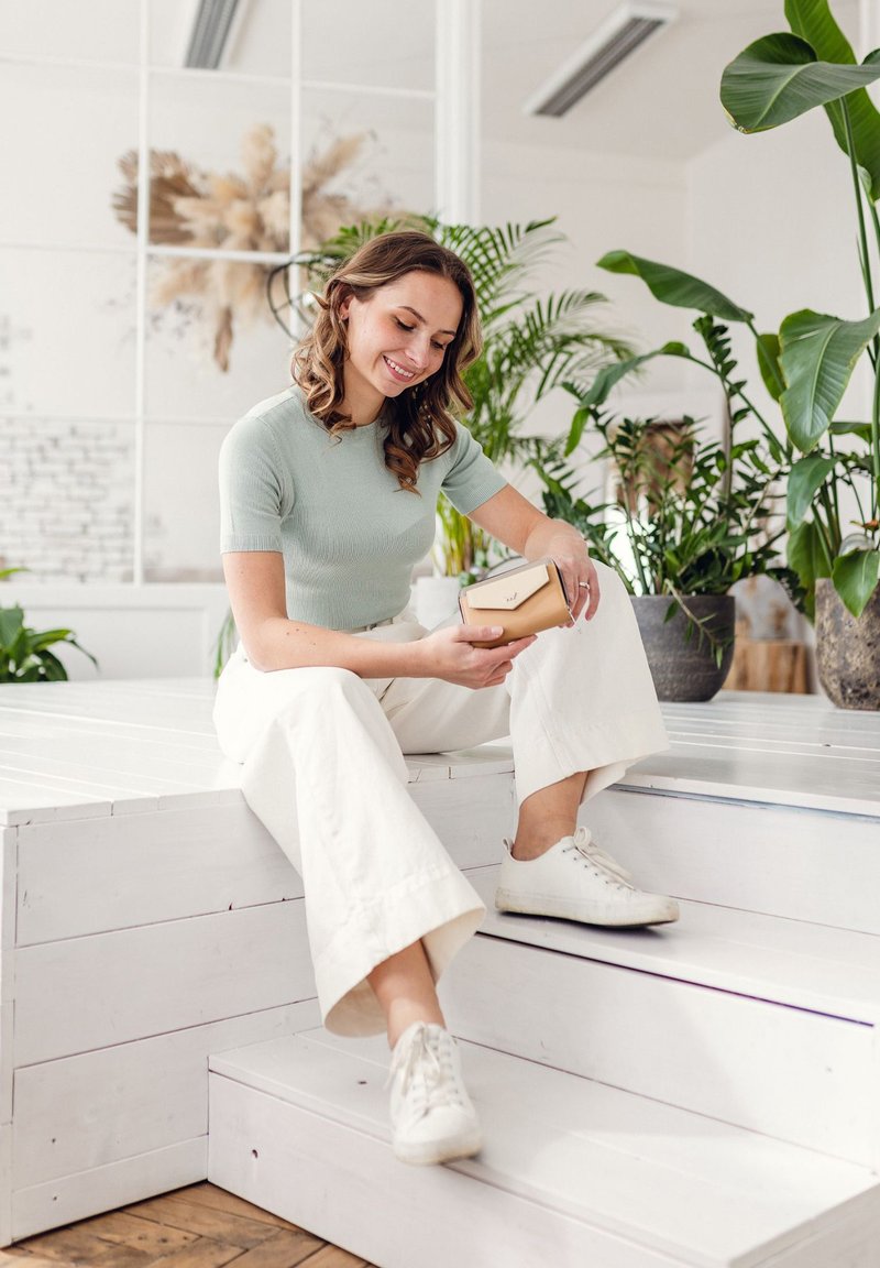 Light green fitted top and cream wide-leg pants; seated on white wooden steps, holding a two-tone wallet amidst indoor plants.