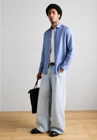 Νέος άνδρας στέκεται σε εσωτερικό χώρο, φορά μπλε πουκάμισο πάνω από λευκό T-shirt, φαρδιά ανοιχτόχρωμα τζιν, μαύρα παπούτσια, μαύρη ζώνη, κρατώντας μια μαύρη τσάντα στο πλάι του.