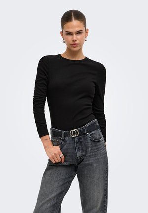 Kvinde med bagstrøget hår iført en sort langærmet top, falmede sorte jeans og et sort bælte med sølv dobbeltrings spænde.