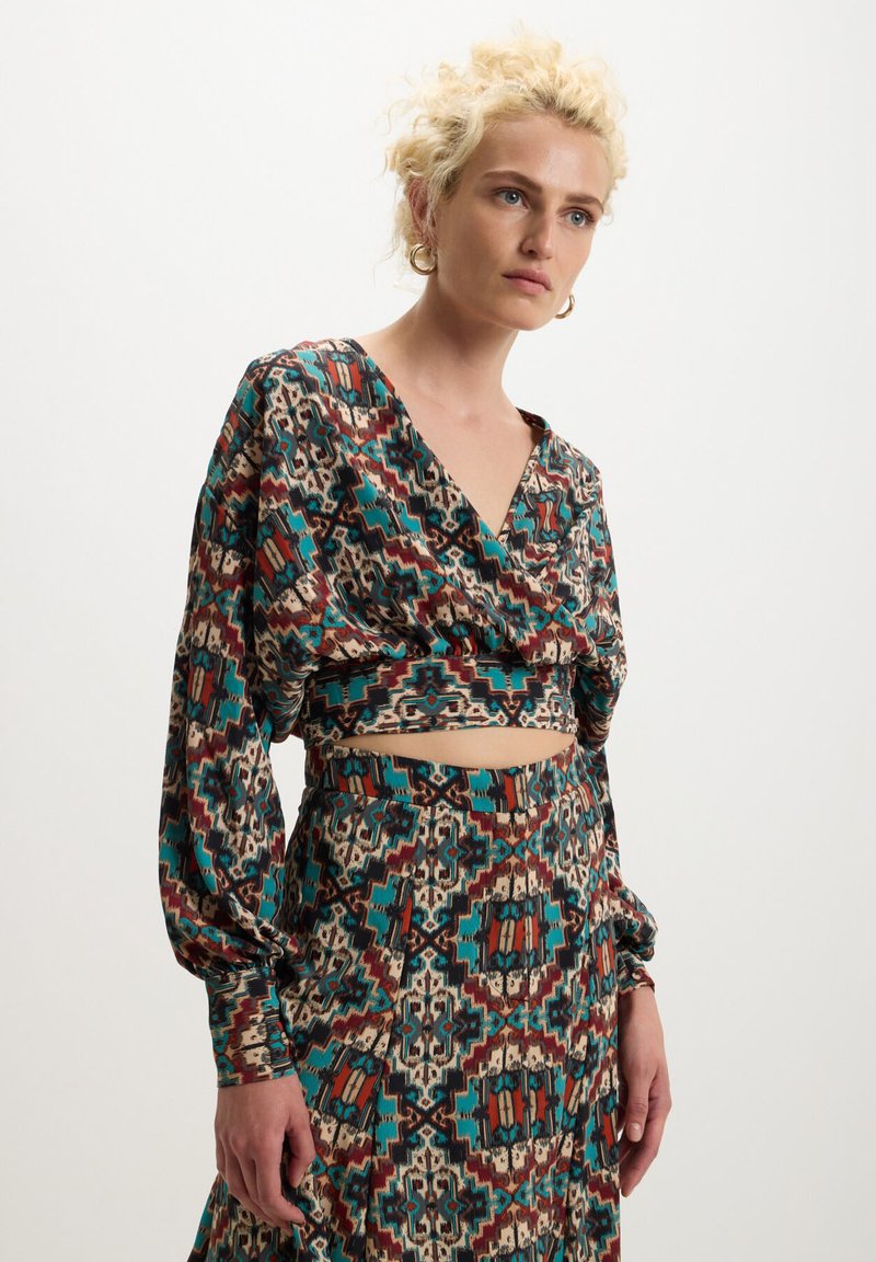 Femme blonde portant des boucles d'oreilles, un crop top à motifs teal, marron et beige, et une jupe assortie, se tenant devant un fond uni.