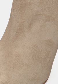 Botín de tobillo de ante beige con una textura suave, forma cónica y un diseño sin costuras con un acabado mate sutil.