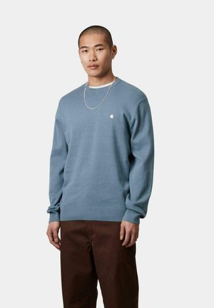 Suéter azul de tela suave, presenta un cuello redondo, puños acanalados y un pequeño logo en el pecho. Combinado con pantalones marrones.
