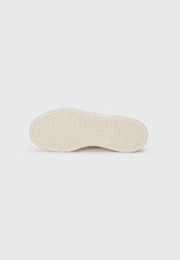 Shoe sole in light beige, featuring a textured pattern with wavy lines and branding. Smooth edges with a slightly raised heel area.