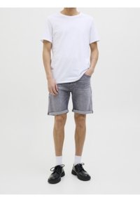 Henkilö seisoo valkoisessa t-paidassa, käärityissä harmaissa farkkushortsissa, valkoisissa sukissa ja mustissa nauhakenkässä tasaisella taustalla.