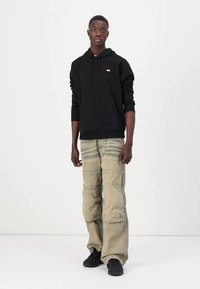 Čierna mikina s kapucňou a predným vreckom, skombinovaná so svetlo hnedými voľnými cargo nohavicami s viacnásobnými švami a čiernymi teniskami.