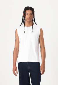 Tricou alb fără mâneci, din bumbac, cu guler rotund și o croială lejeră, asortat cu blugi negri din denim.